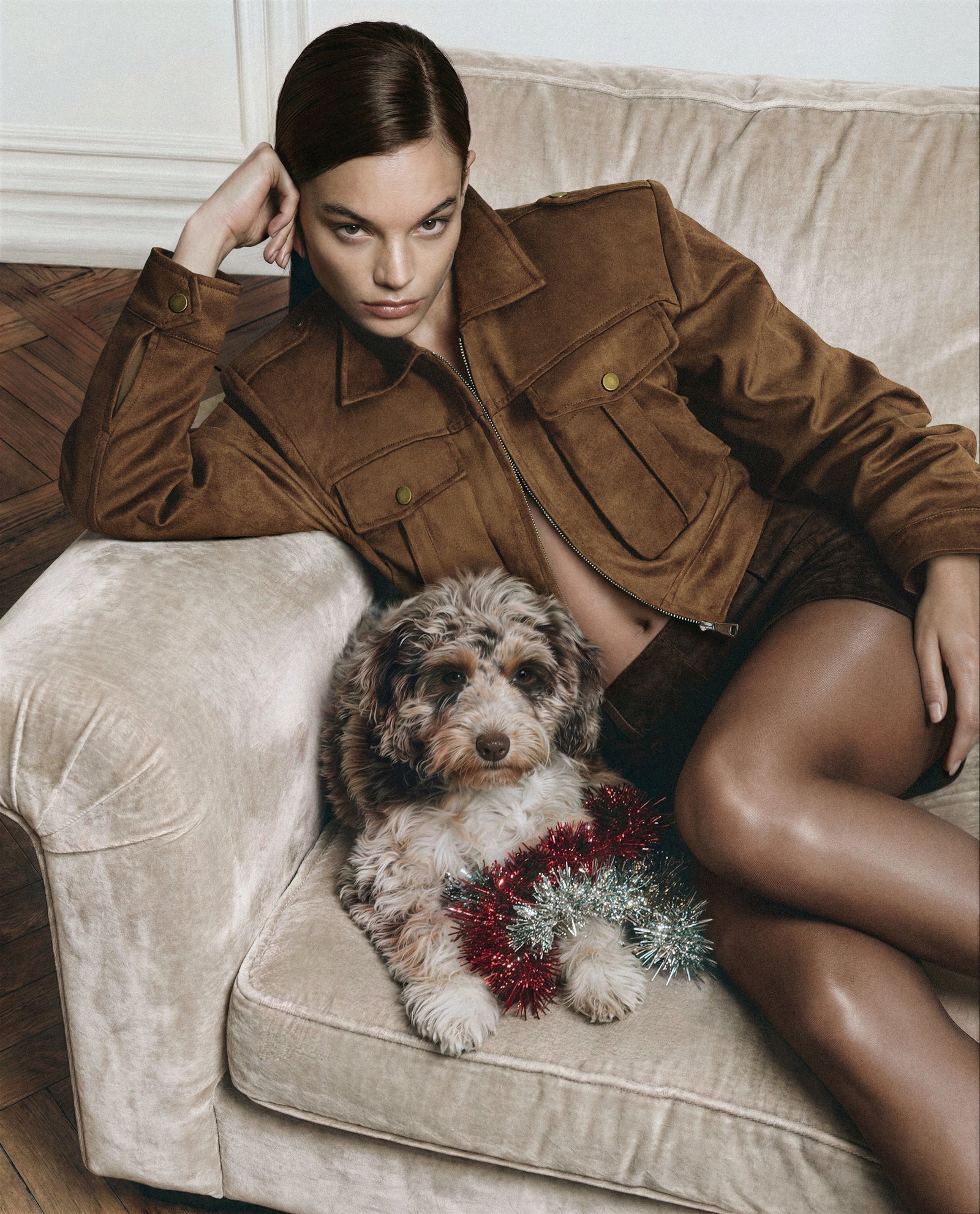 Woman in a brown jacket sitting on a beige couch with a small dog wearing a red and silver sweater.